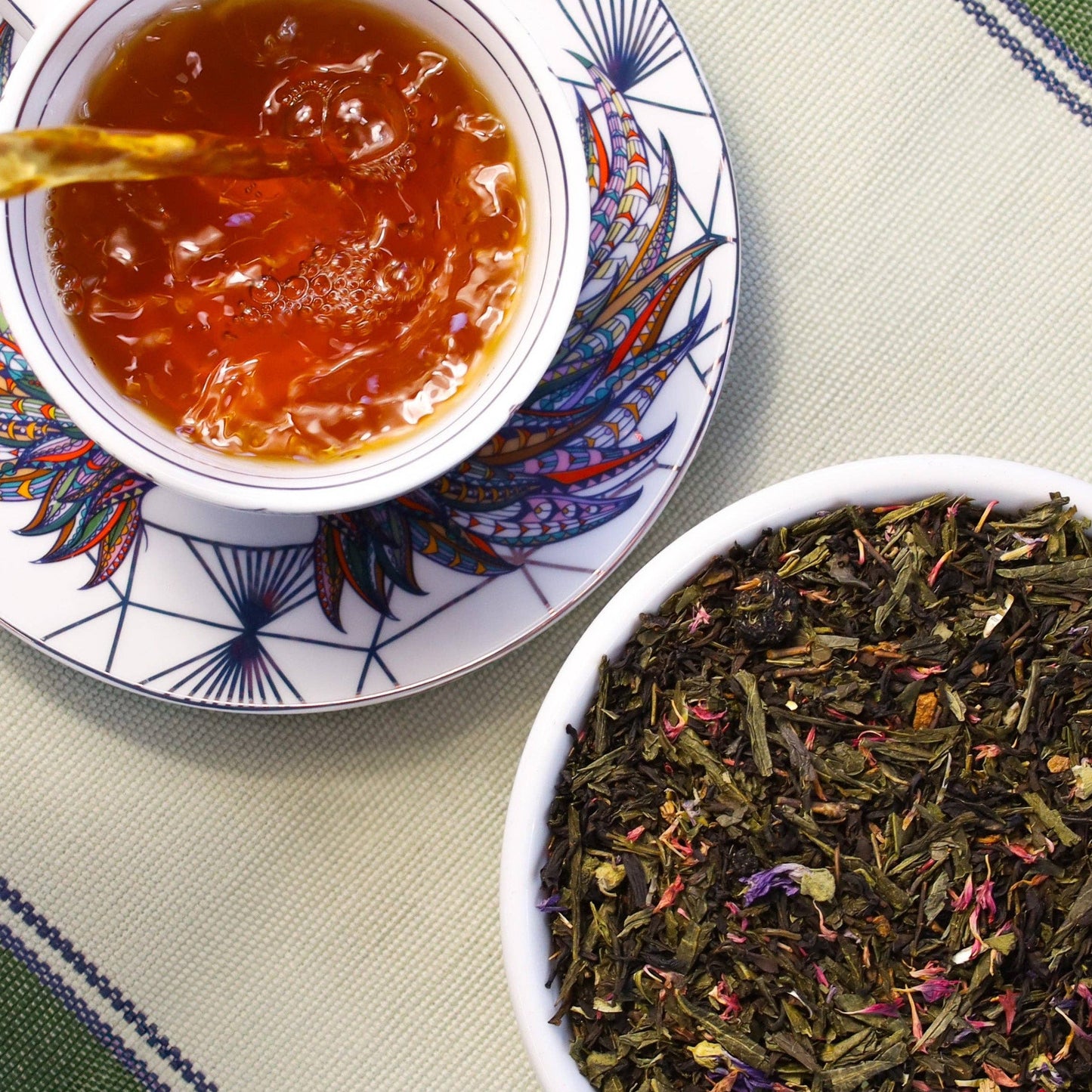 A top-down view of a white teacup and saucer filled with amber tea, alongside a white bowl of loose-leaf herbal tea blend on a patterned table mat.