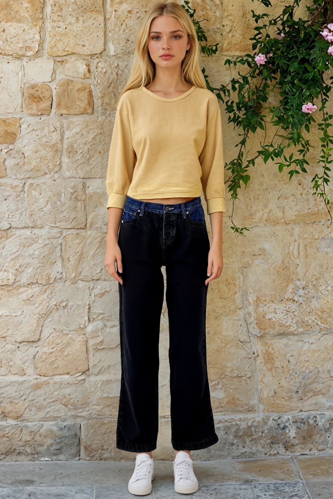 Woman wearing a yellow sweater and black pants standing against a stone wall with greenery.