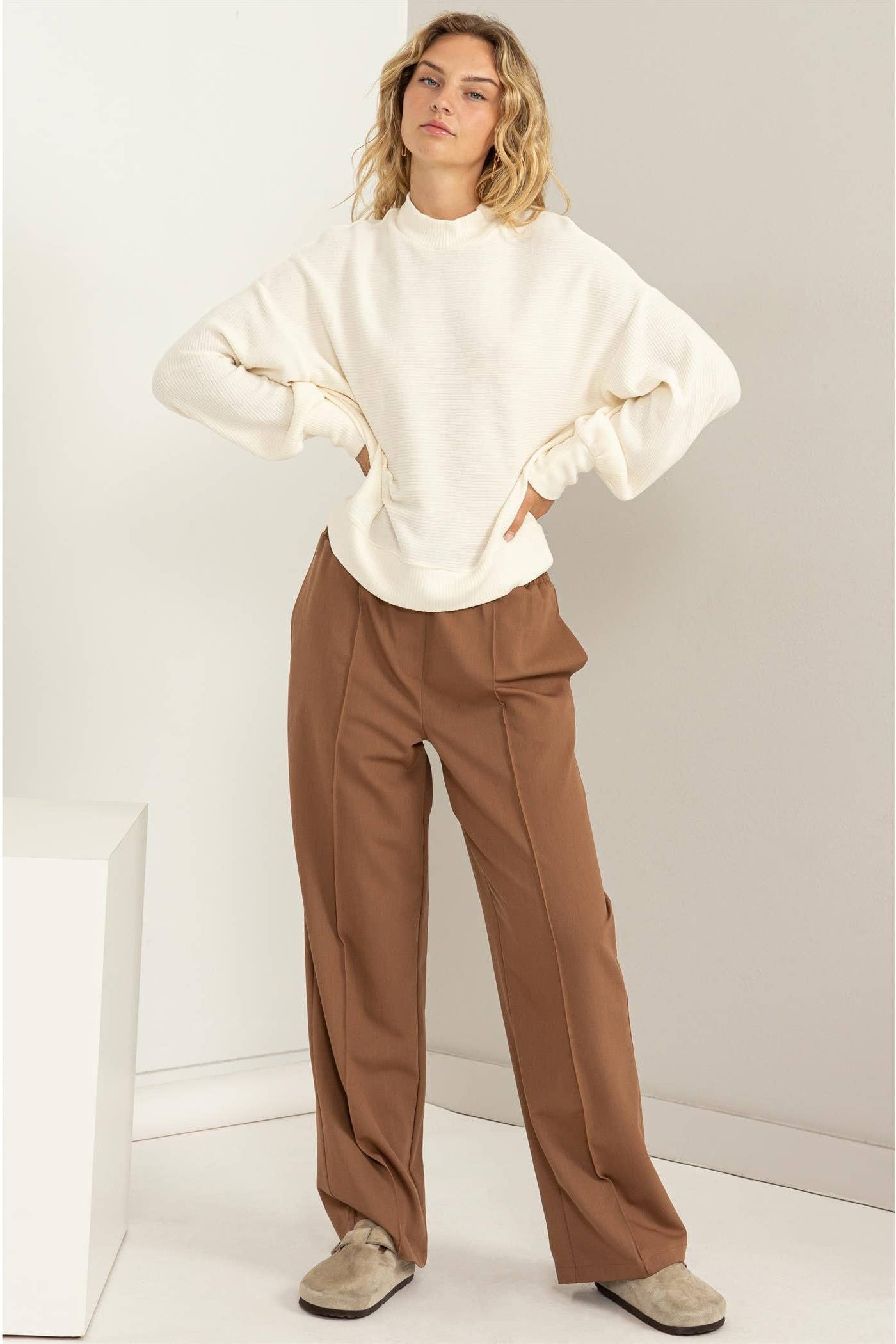 Woman wearing a white sweater and brown pants against a white background