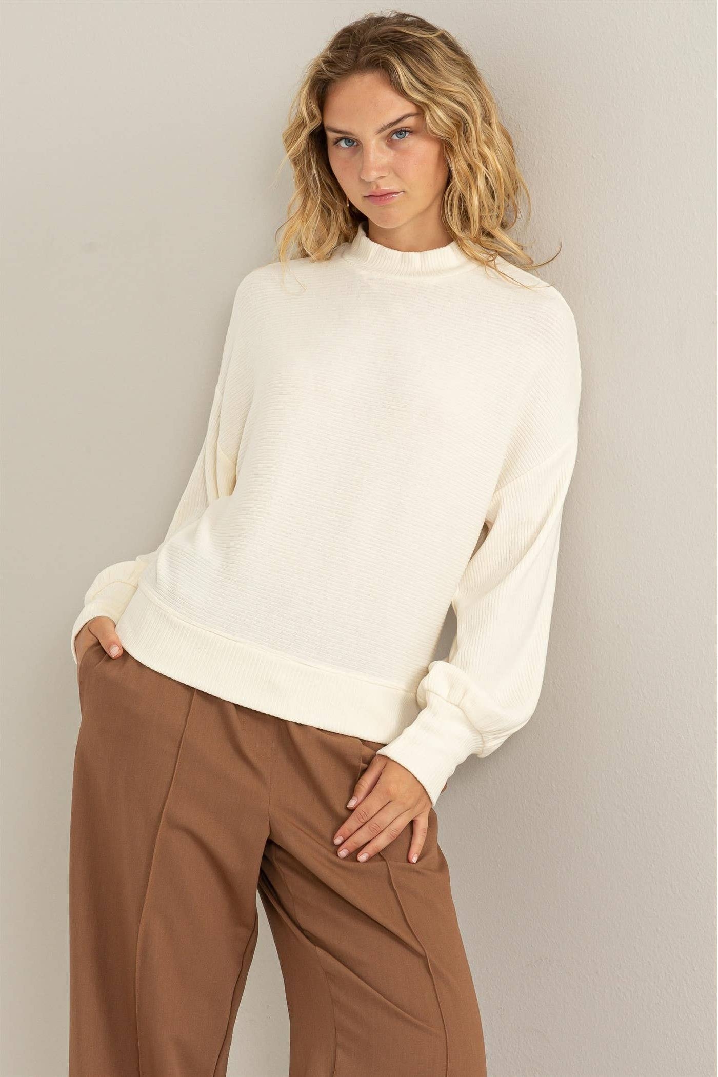 Woman wearing a white sweater and brown pants against a plain background