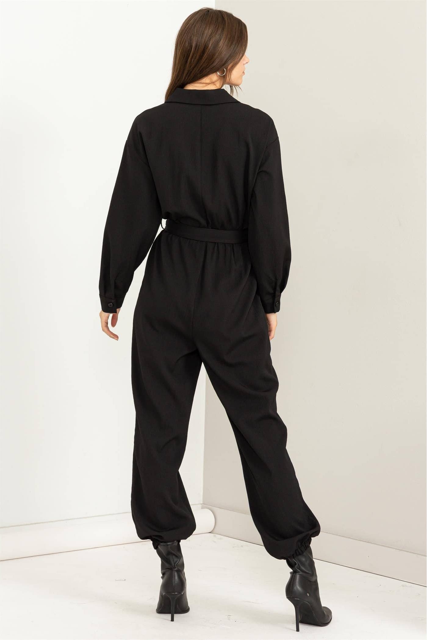 Person wearing a black jumpsuit standing against a white wall.