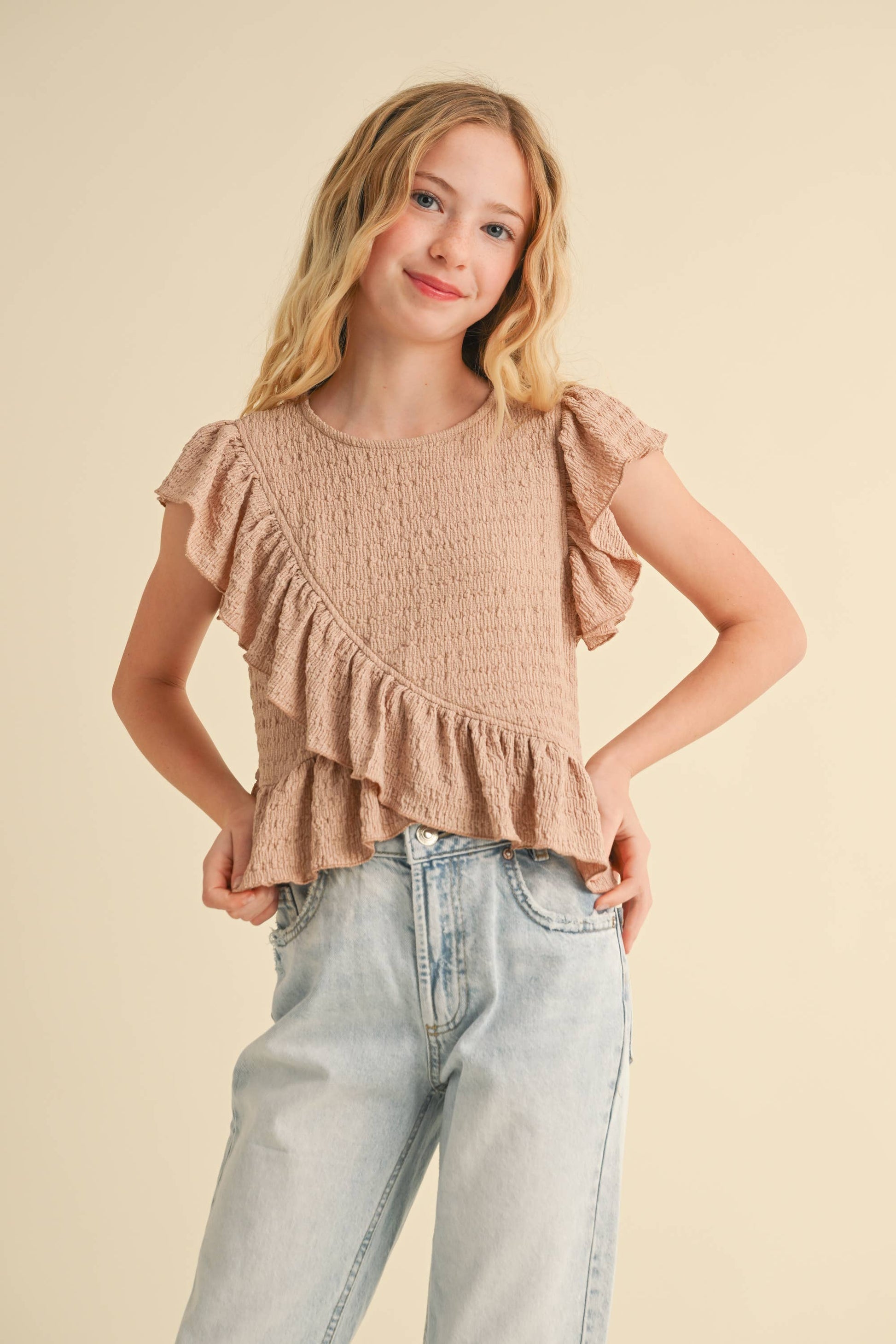 Young girl wearing a beige ruffled top and light blue jeans on a beige background