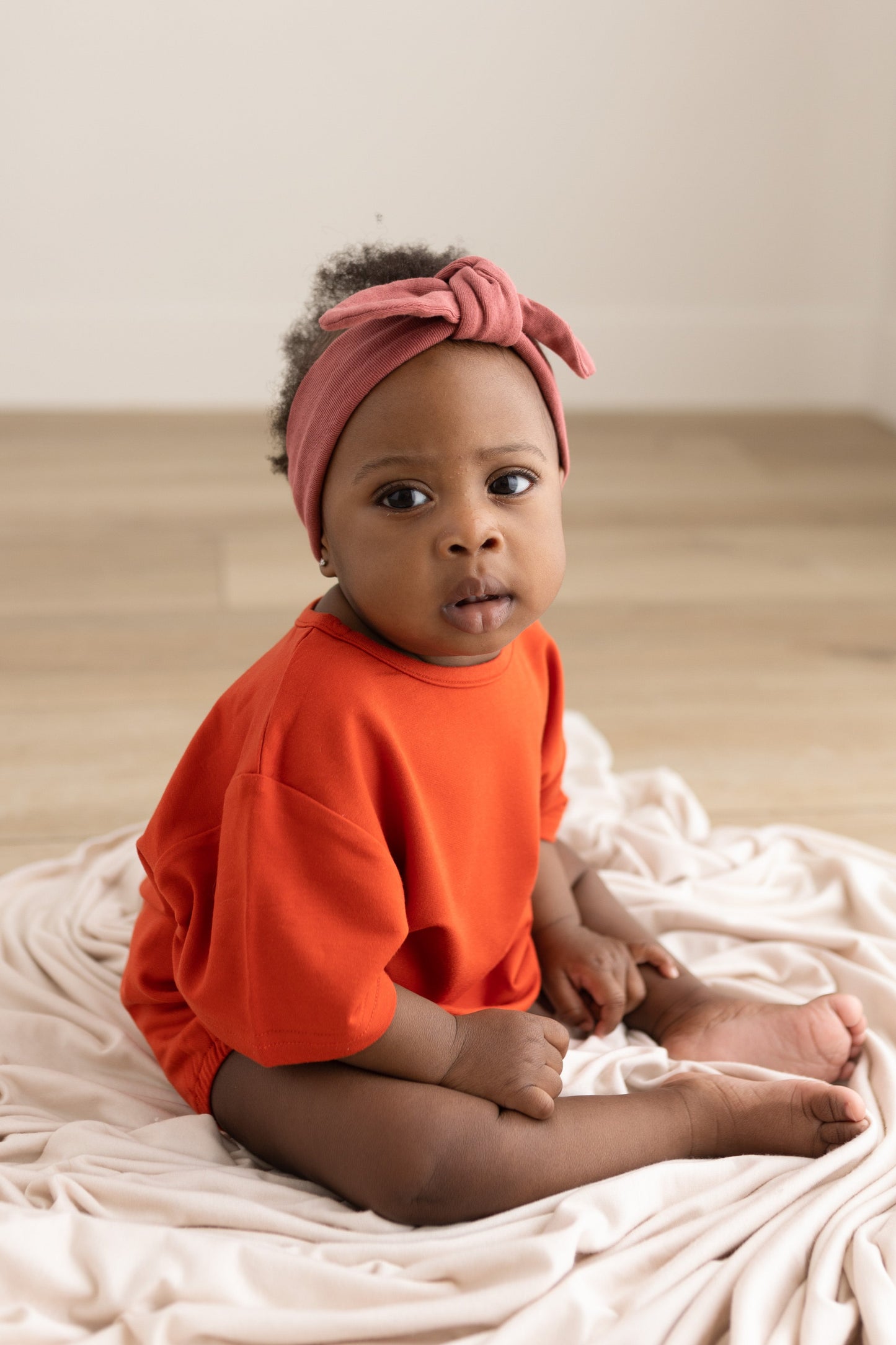 Baby wearing an orange shirt and pink headband sitting on a white blanket.