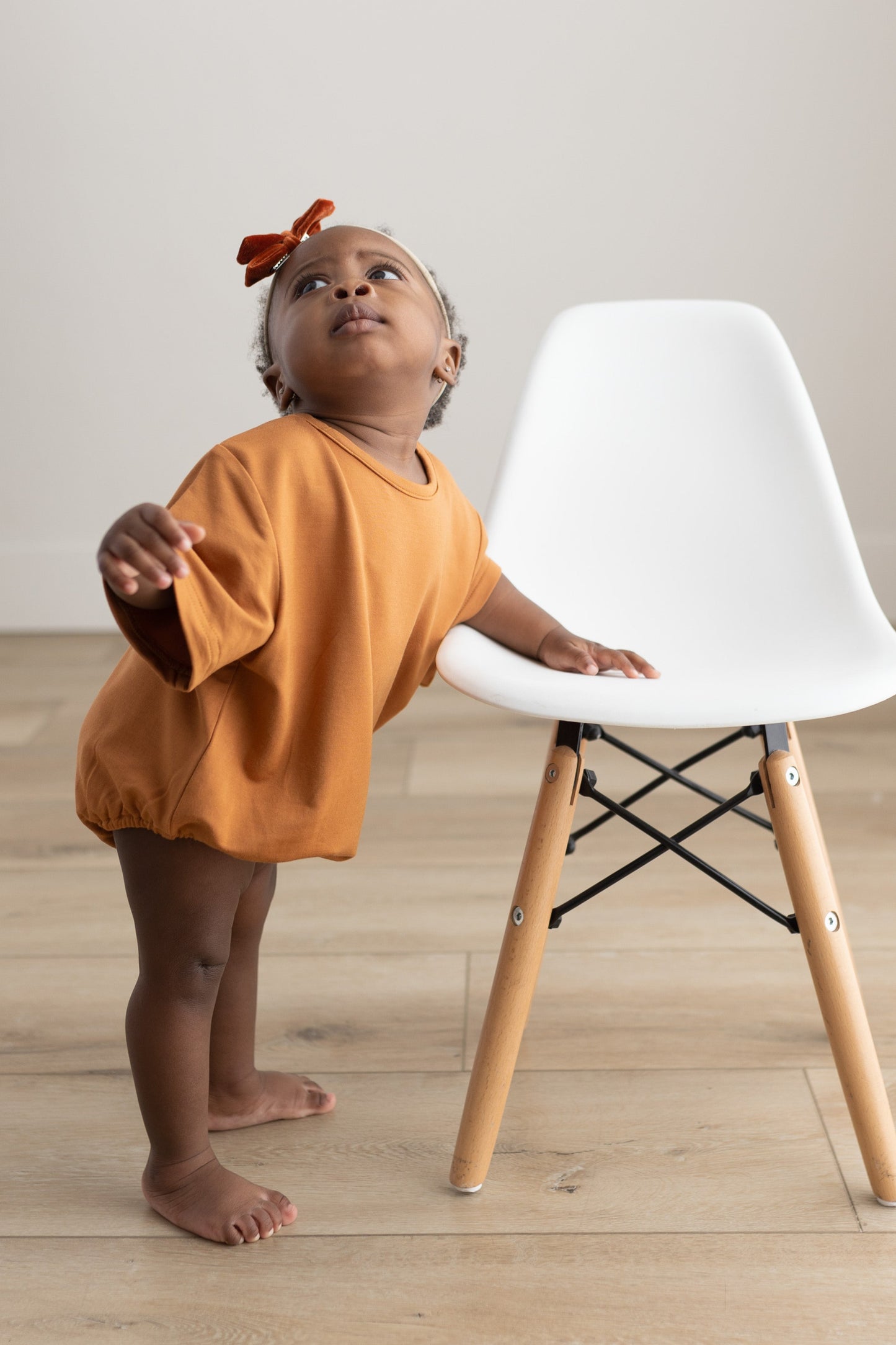 Child in an orange outfit standing next to a white chair with wooden legs on a light wood floor.