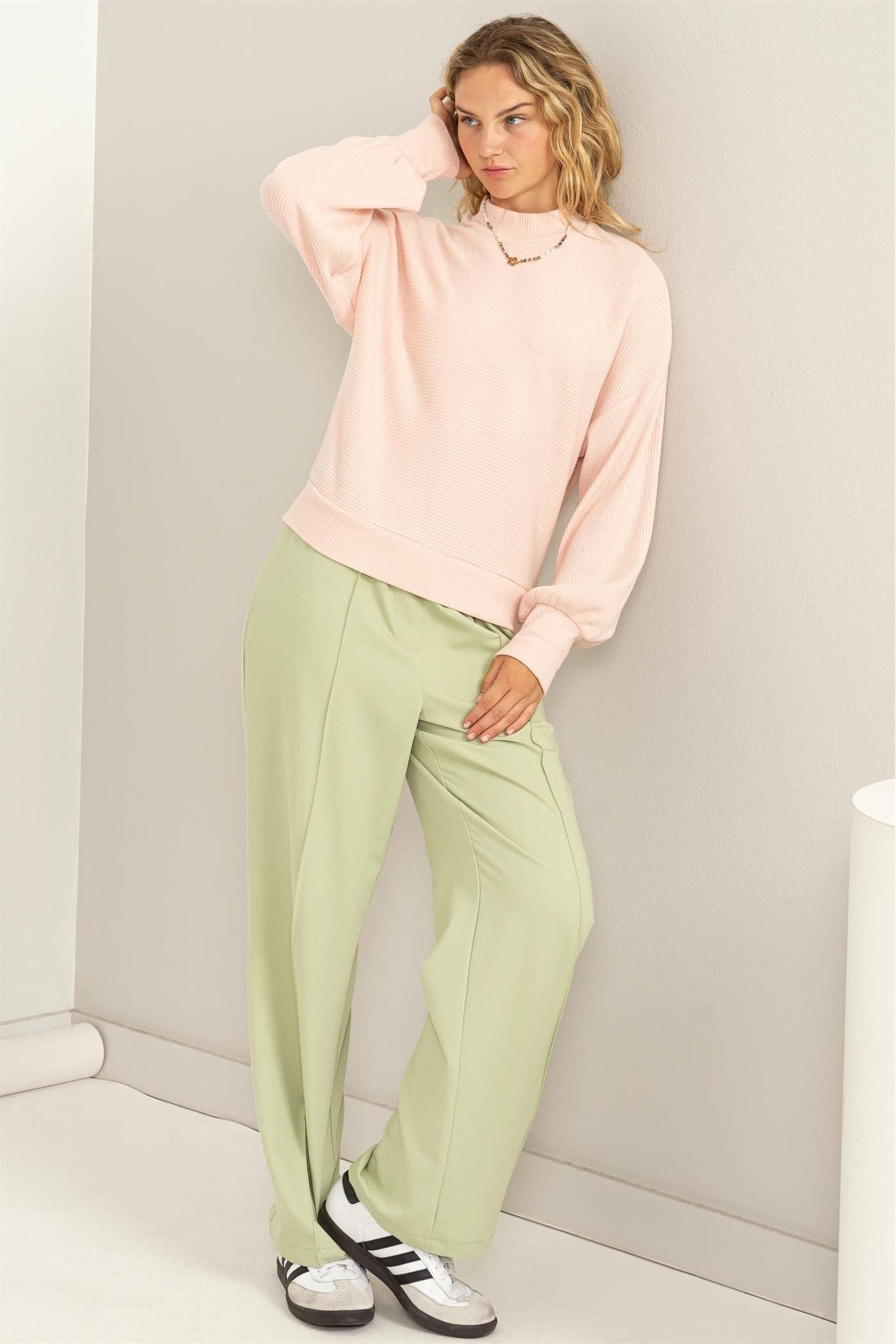 Woman wearing a light pink sweater and green pants against a plain background