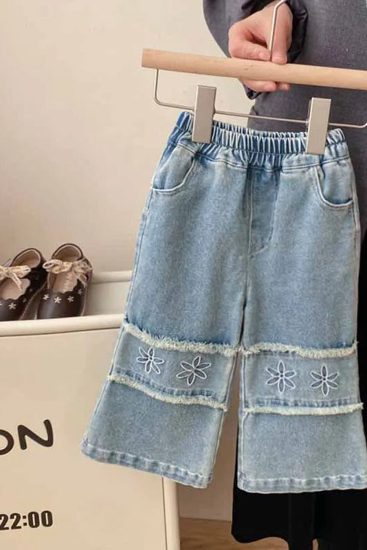 Denim pants with floral embroidery on a hanger, with shoes and a bag in the background.