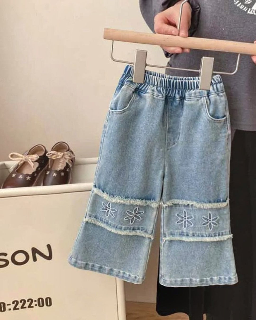 Denim pants with floral embroidery on a hanger, with shoes and a bag in the background.