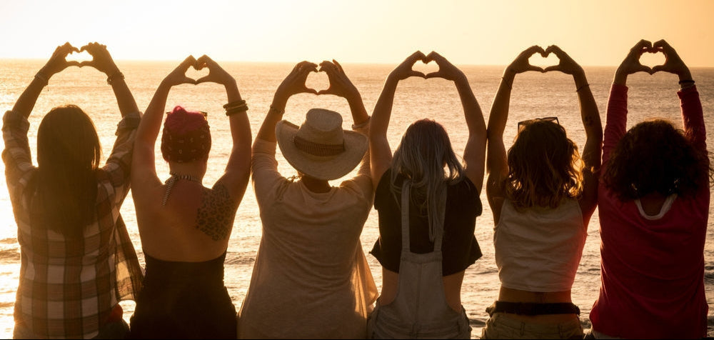 Group of people sitting on a beach at sunset, making heart shapes with their hands.