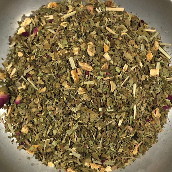 A close-up top-down view of loose-leaf herbal tea mixed with dried botanicals, including green leaves, light tan sticks, and small purple flower pieces, piled in a clear container.