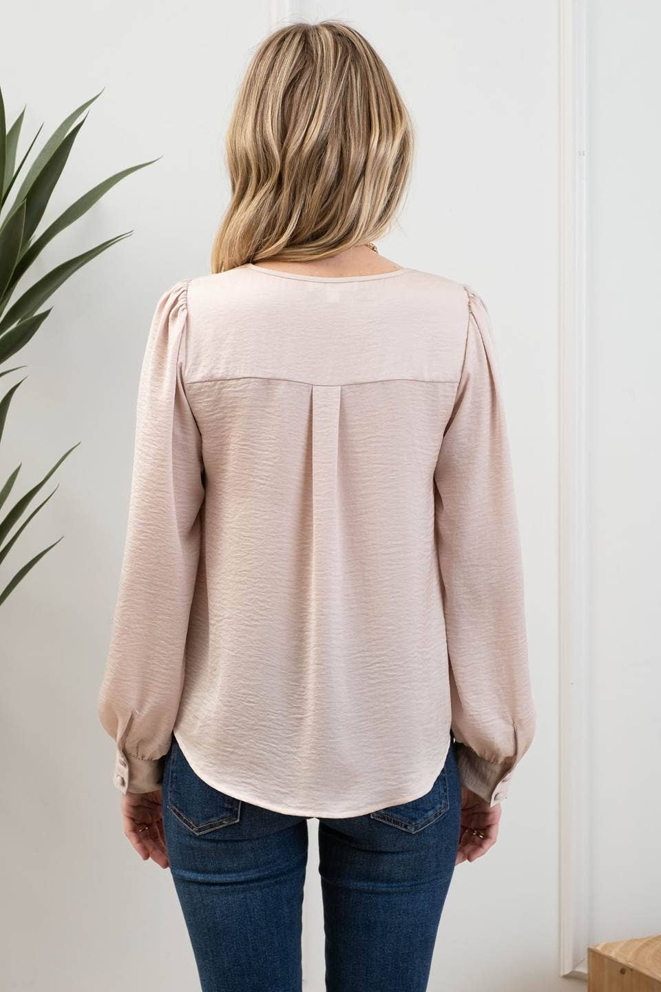 Person wearing a beige blouse and blue jeans standing against a white wall with a plant to the left.