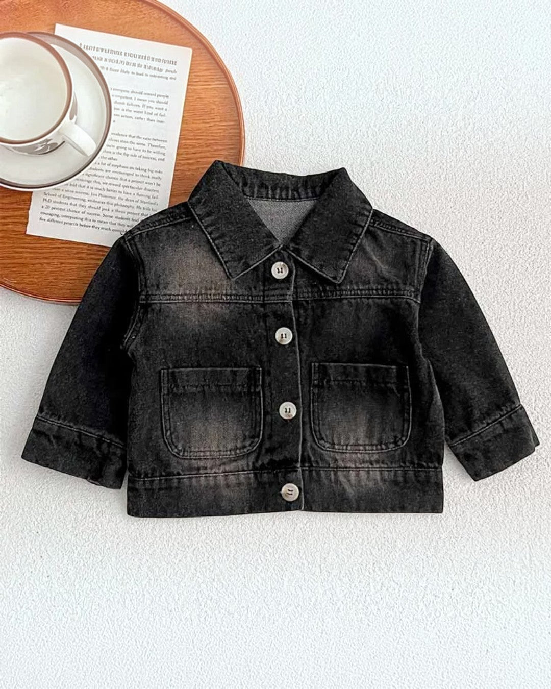 Black denim jacket on a white surface with a wooden coaster and saucer in the background.