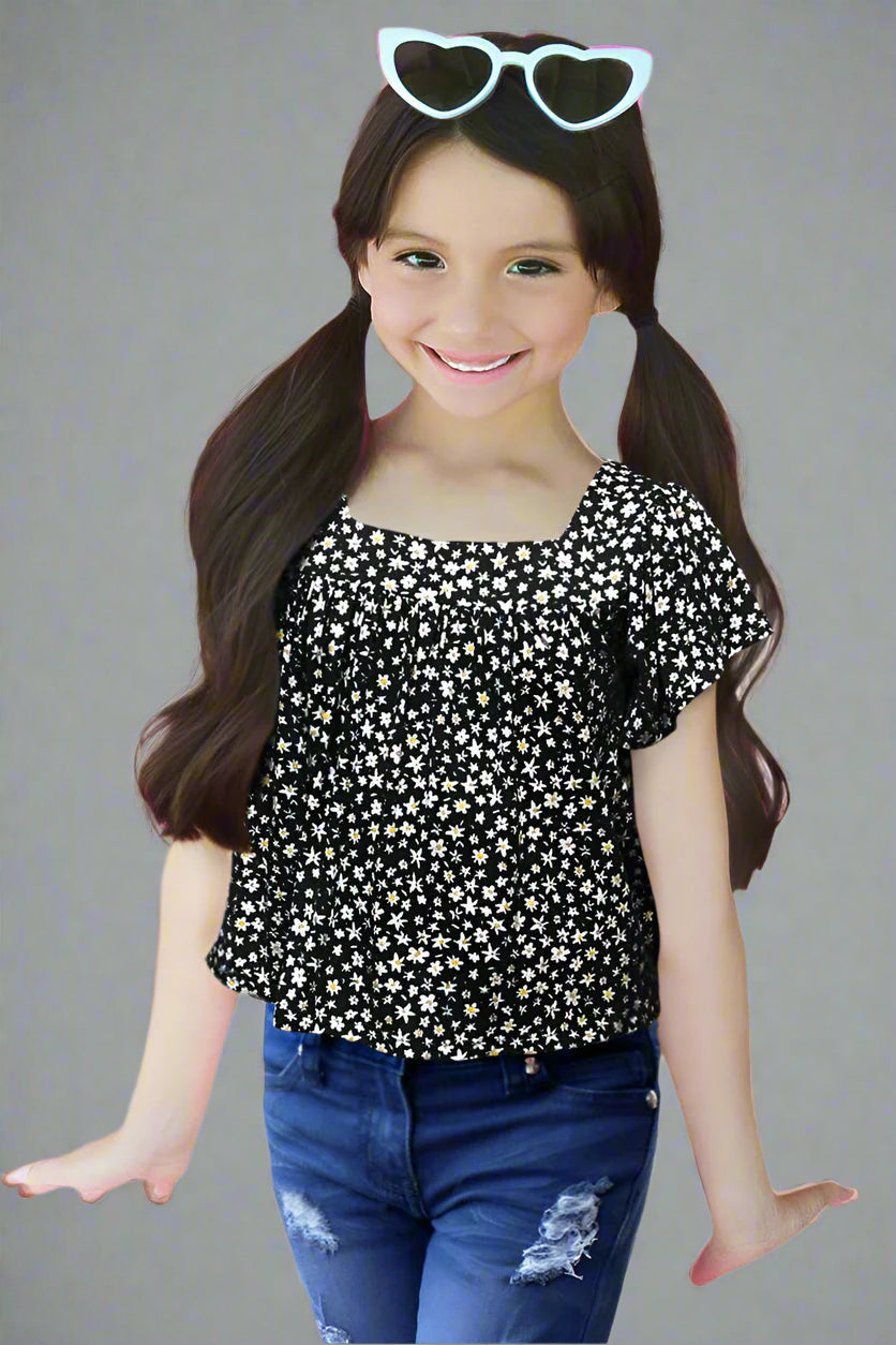 Young girl wearing a black floral top and blue jeans with heart-shaped sunglasses on her head against a plain background.