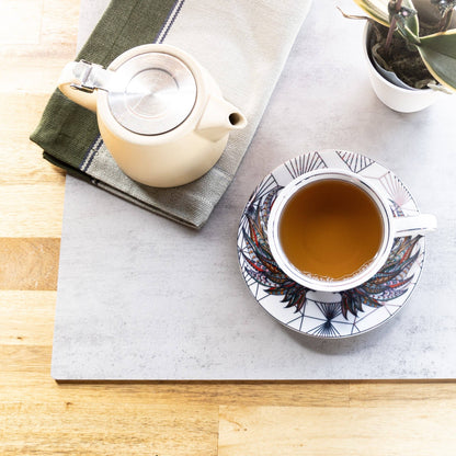 A ceramic teapot with a silver lid sits on a folded green and gray striped cloth, next to a white teacup and matching saucer filled with tea, arranged on a gray tray over a light wood surface.