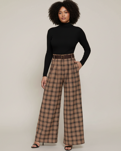 A model with curly black hair stands against a light beige studio background wearing a black long-sleeve mock-neck top and high-waisted brown plaid wide-leg trousers with a matching brown belt.