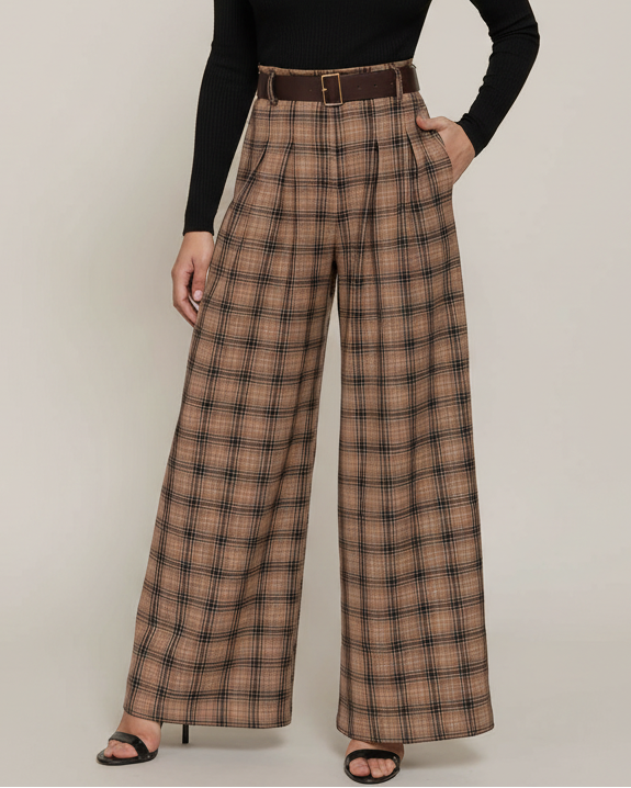 A model with curly black hair stands against a light beige studio background wearing a black long-sleeve mock-neck top and high-waisted brown plaid wide-leg trousers with a matching brown belt.