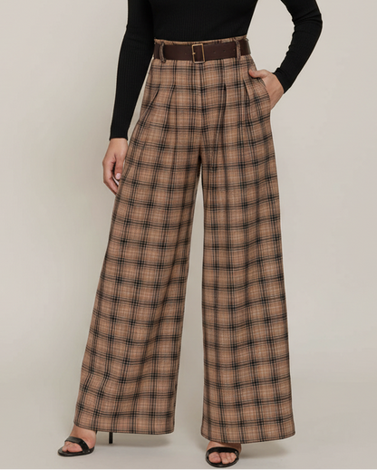 A model with curly black hair stands against a light beige studio background wearing a black long-sleeve mock-neck top and high-waisted brown plaid wide-leg trousers with a matching brown belt.