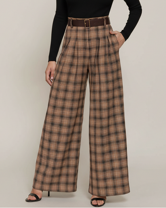 A model with curly black hair stands against a light beige studio background wearing a black long-sleeve mock-neck top and high-waisted brown plaid wide-leg trousers with a matching brown belt.