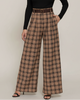 A model with curly black hair stands against a light beige studio background wearing a black long-sleeve mock-neck top and high-waisted brown plaid wide-leg trousers with a matching brown belt.
