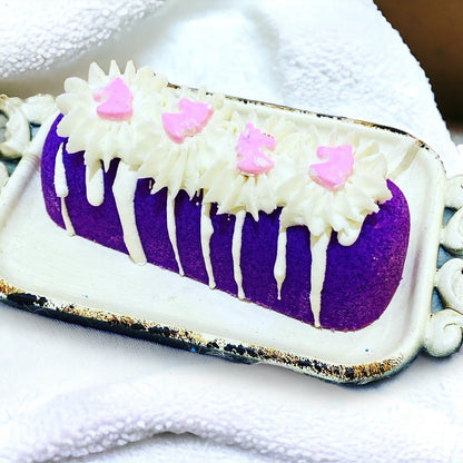 A purple twinkie bath bomb topped with white frosting and light purple sugar sprinkles on a white tray on a white towel