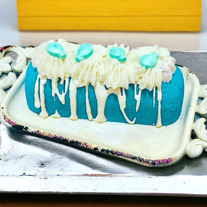 A teal twinkie bath bomb topped with white frosting and teal sugar sprinkles on a white tray on top of a white countertop