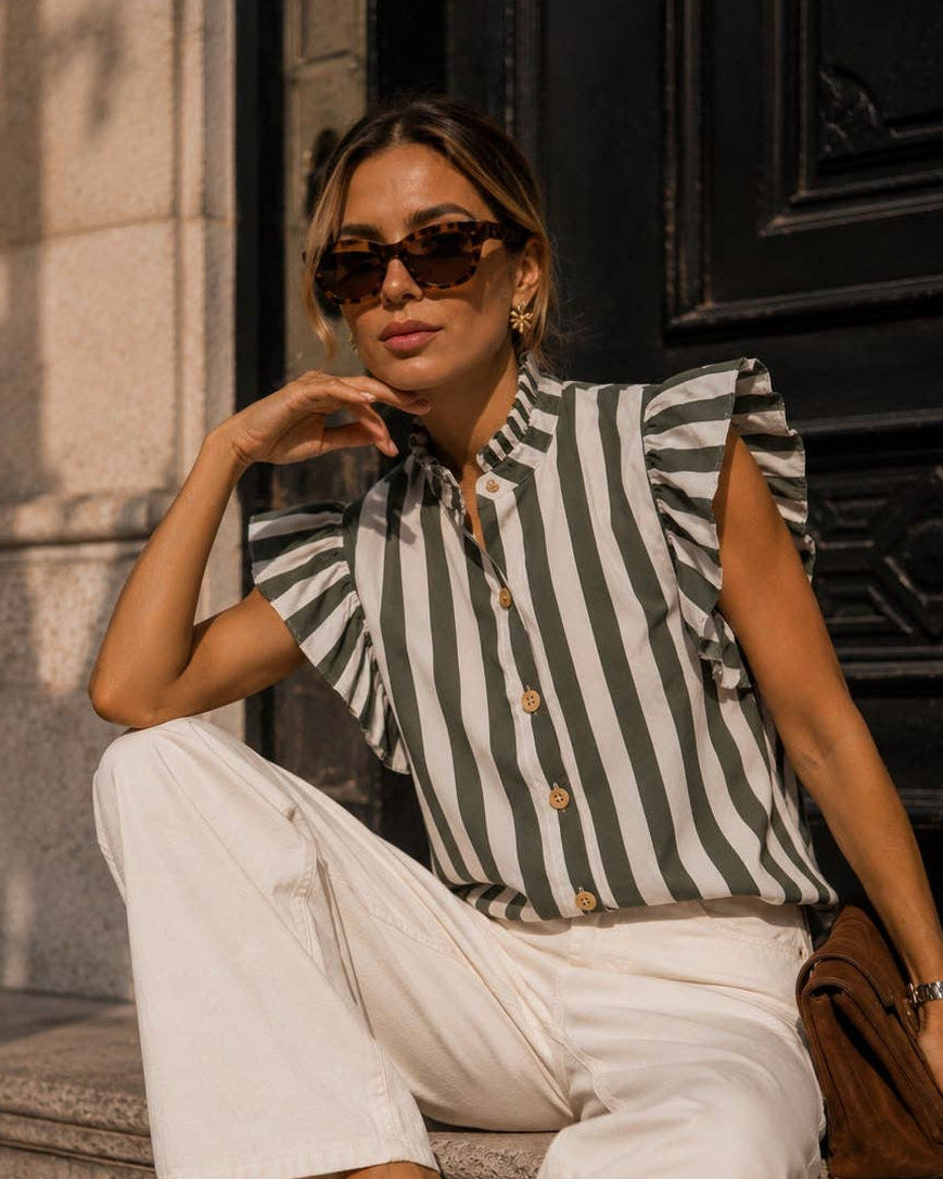 A woman sits on stone steps wearing a black and white vertically striped short-sleeve blouse with ruffled shoulders, white wide-leg pants, brown flat shoes, and sunglasses, holding a brown handbag.