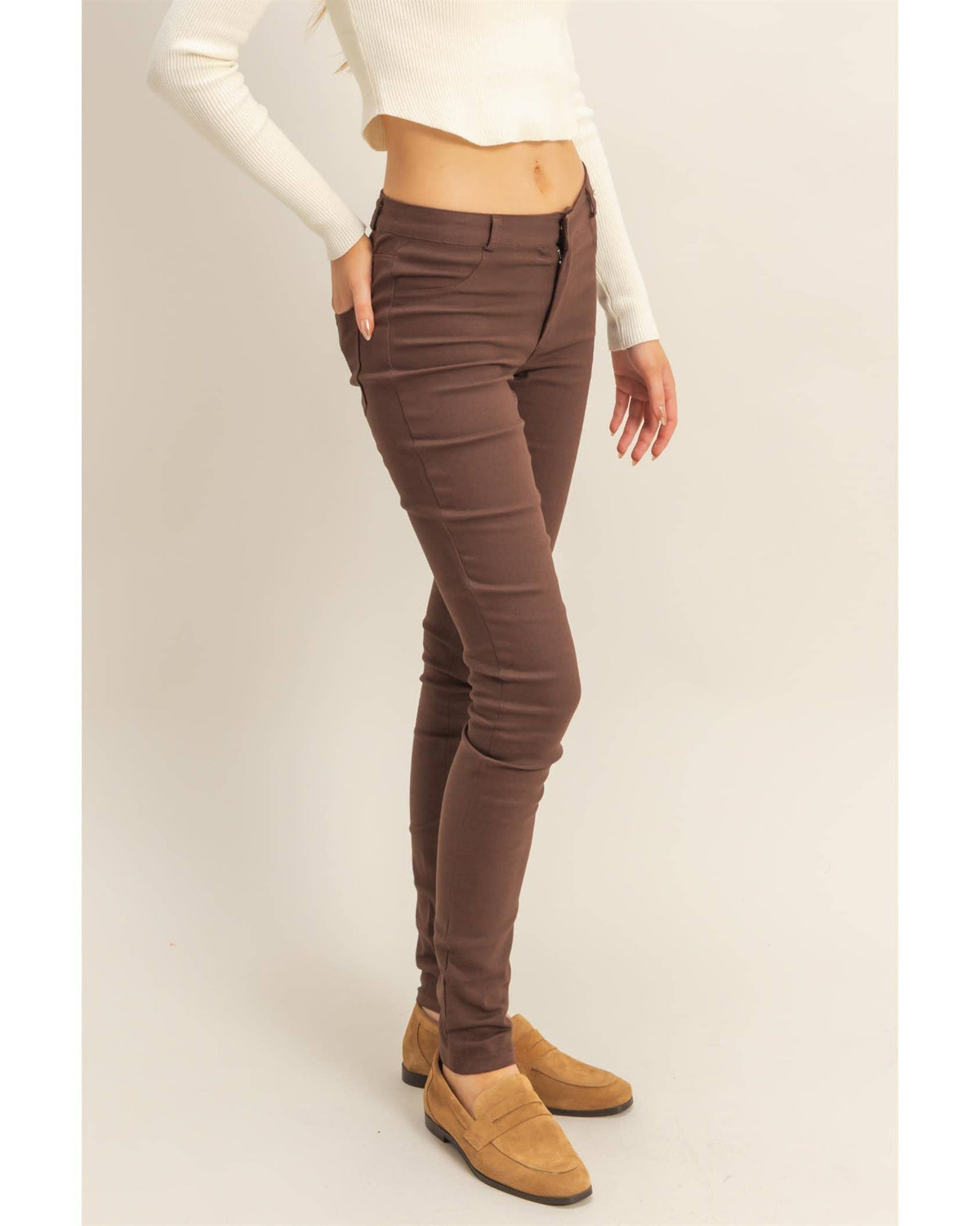 A model wearing slim-fit brown skinny jeans, styled with a cream long-sleeve crop top and tan suede loafers, standing against a plain beige studio background.