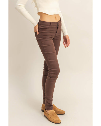 A model wearing slim-fit brown skinny jeans, styled with a cream long-sleeve crop top and tan suede loafers, standing against a plain beige studio background.