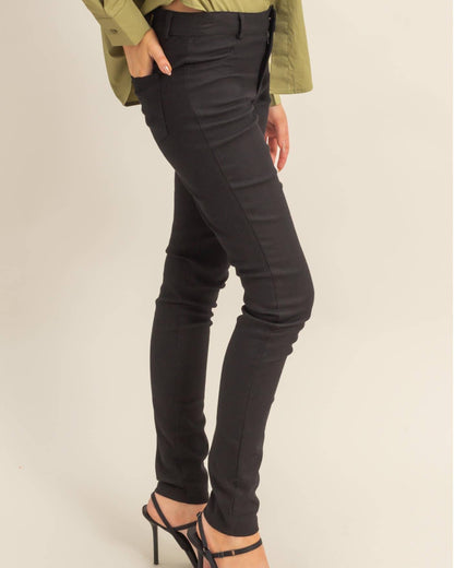 A model wearing slim-fit black skinny jeans, styled with an olive green long-sleeve shirt and black strappy high-heeled sandals, shown from the waist down against a plain beige studio background.