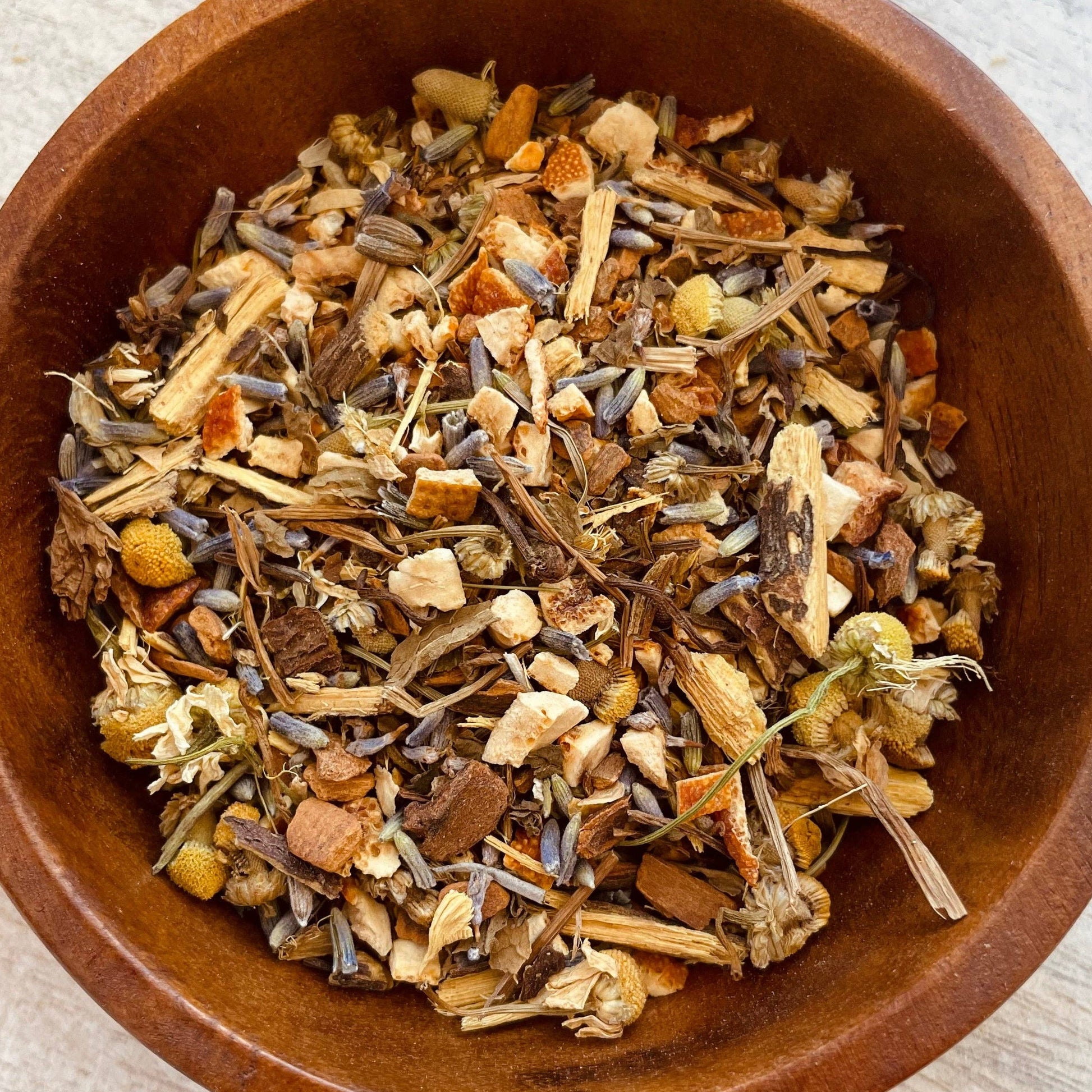 A top-down view of a loose herbal tea blend inside a round wooden bowl, set on a light-colored wooden surface.