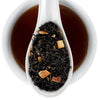 A white ceramic spoon filled with loose-leaf tea is centered over a white cup of dark brewed tea on a white background. The brand name 'Finest Elmwood County Inn' is printed in the top right corner.