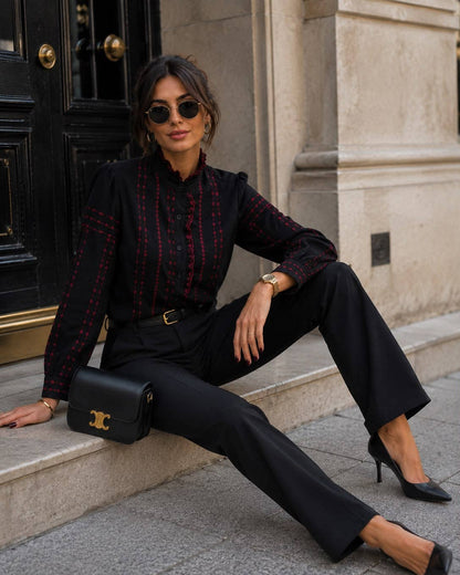 A woman sits on stone steps wearing a black long-sleeve button-up shirt with red and gold plaid details, black wide-leg trousers, black pointed heels, and a black shoulder bag with gold hardware.