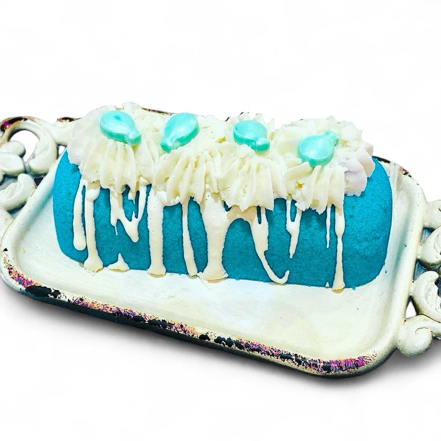 A teal twinkie bath bomb topped with white frosting and teal sugar sprinkles on a white tray against a white background