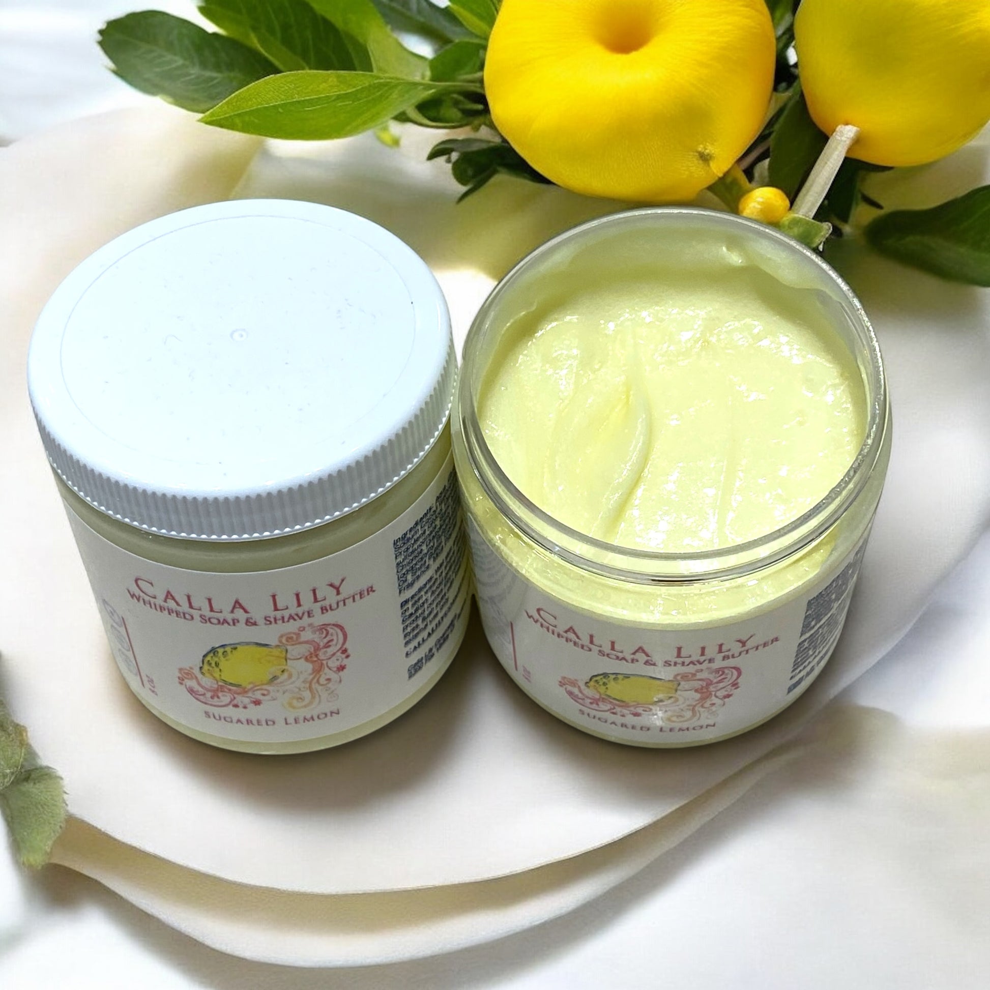 Two jars of 'Calla Lily Whipped Soap & Shave Butter' with lemon slices and leaves on a white plate.