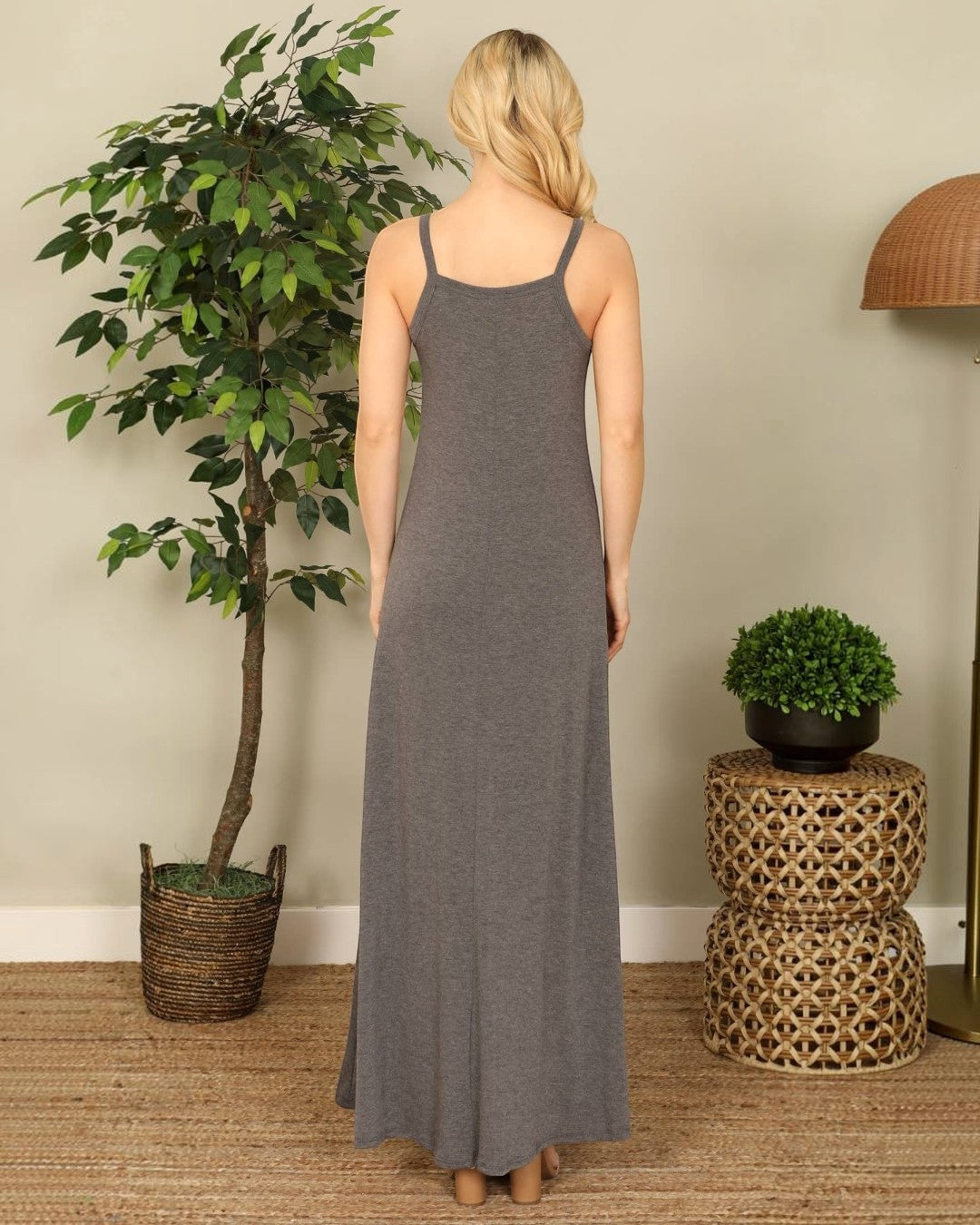 Woman wearing a long gray dress standing in a room with plants and a woven side table.