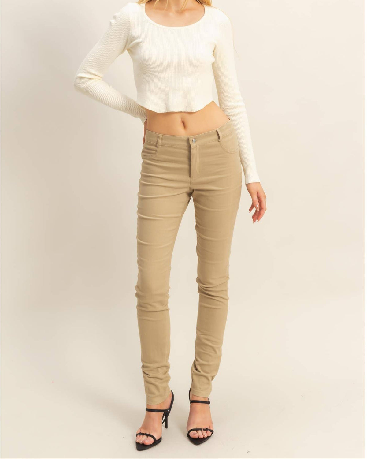 A woman modeling a fitted cream long-sleeve crop top and beige skinny jeans with black high-heeled sandals, standing against a plain light beige studio background.
