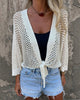 Person wearing a white crochet cardigan over a black top and denim shorts against a stone wall.