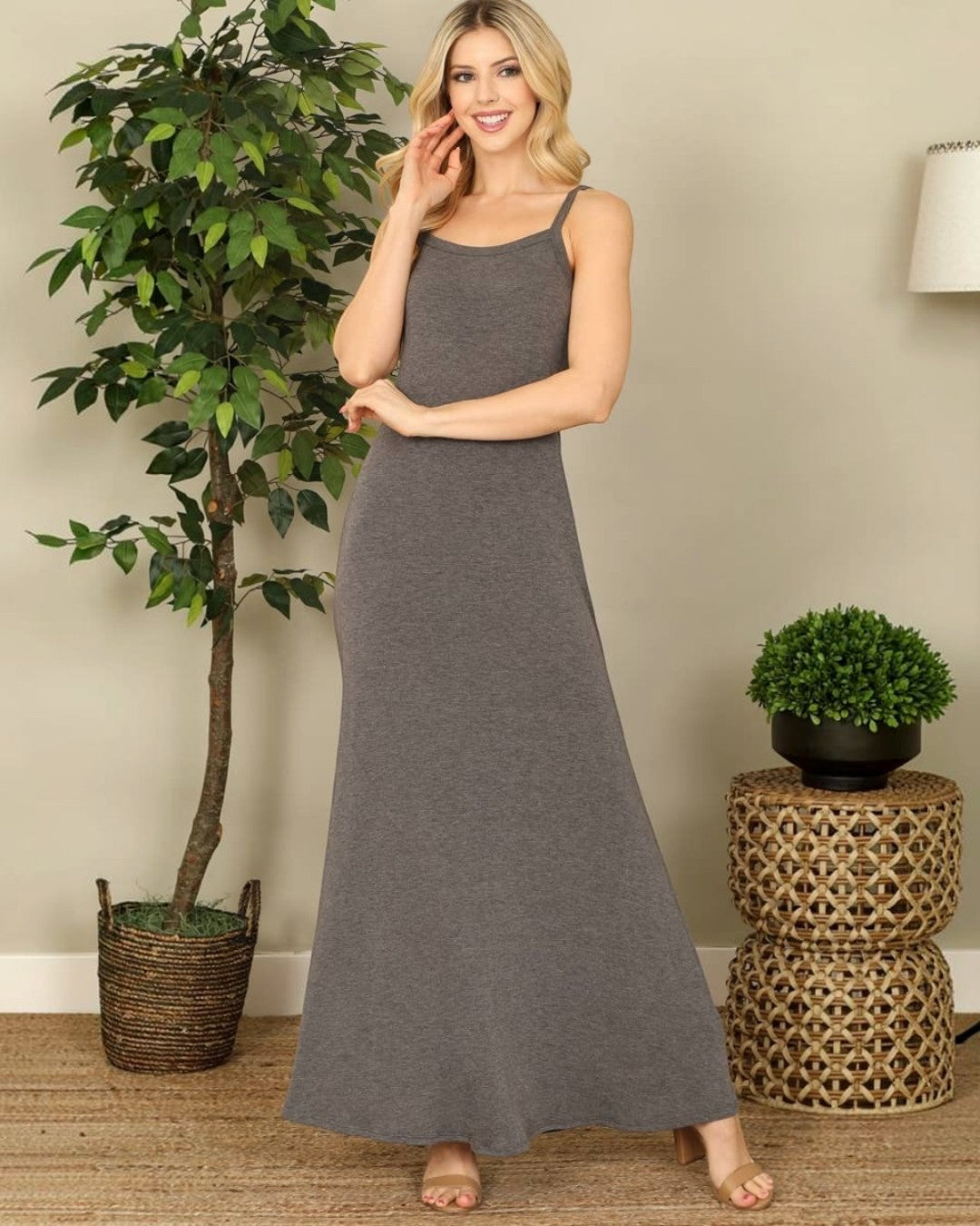 Woman wearing a gray dress standing in a room with plants and a woven stool.