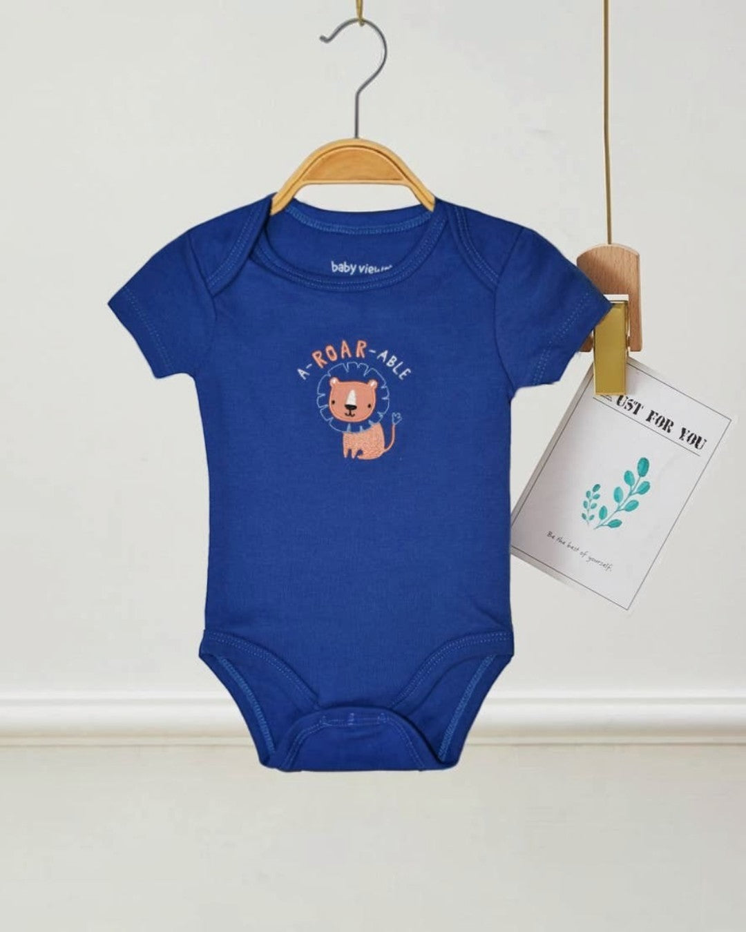 Blue baby onesie with a lion graphic and text on a hanger against a white wall.