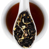 A white ceramic spoon resting over a white cup filled with dark amber tea, with loose-leaf tea and dried flower petals visible in the spoon. The Fine Elmwood Inn logo appears in the top right corner.