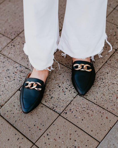 Black loafers with gold chain detail worn on a stone pavement