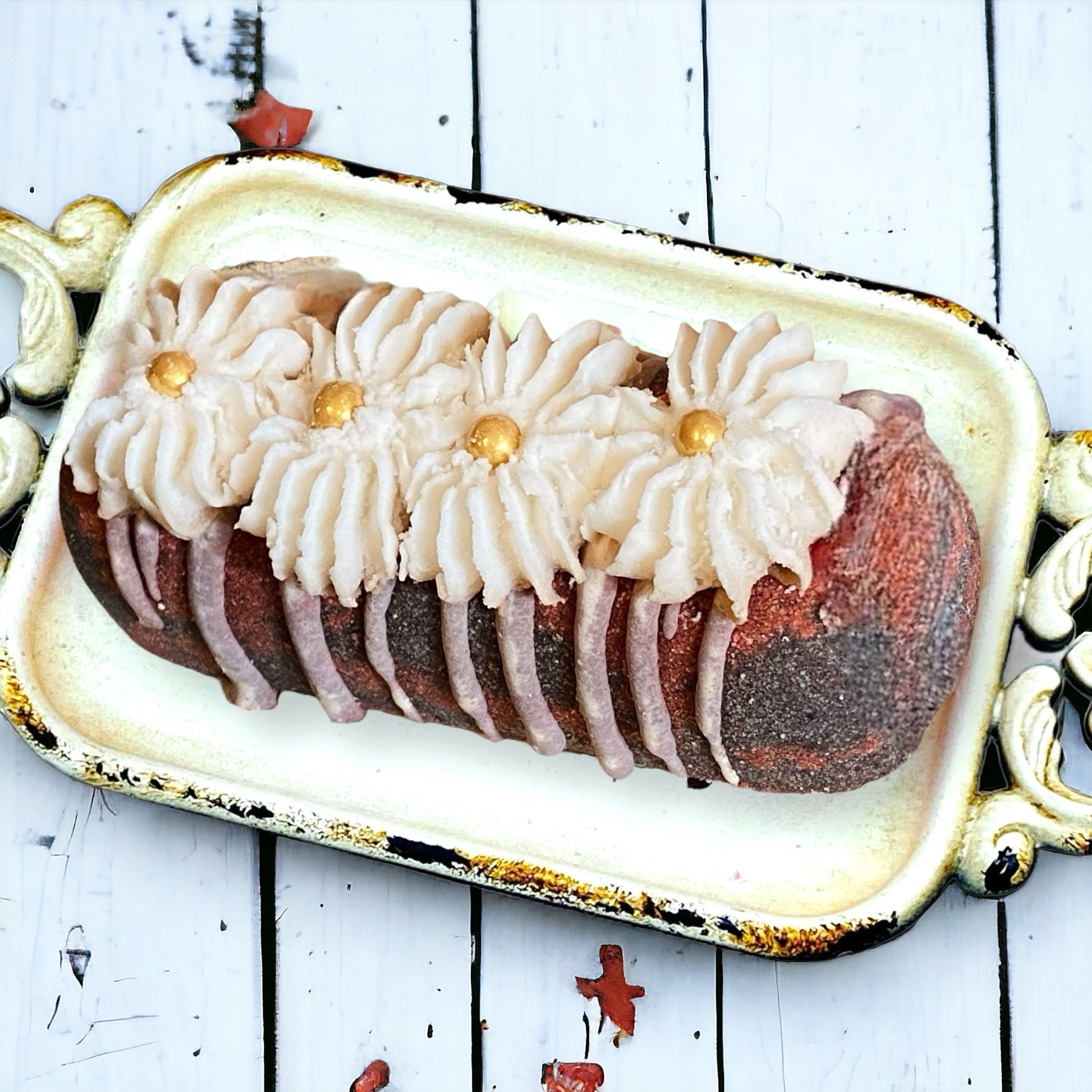 A red and black twinkie bath bomb topped with white frosting and gold sugar sprinkles on a white tray against a wood background