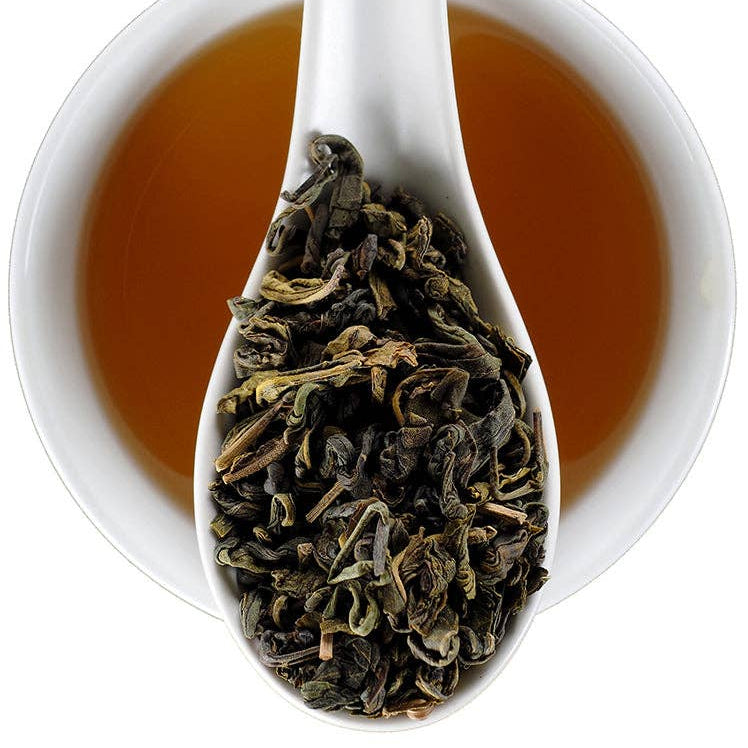 A white ceramic cup filled with amber tea, with a white spoon resting inside the cup and holding loose dark green tea leaves. The Elmwood Inn logo is visible in the top right corner.