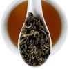 A white ceramic cup filled with amber tea, with a white spoon resting inside the cup and holding loose dark green tea leaves. The Elmwood Inn logo is visible in the top right corner.