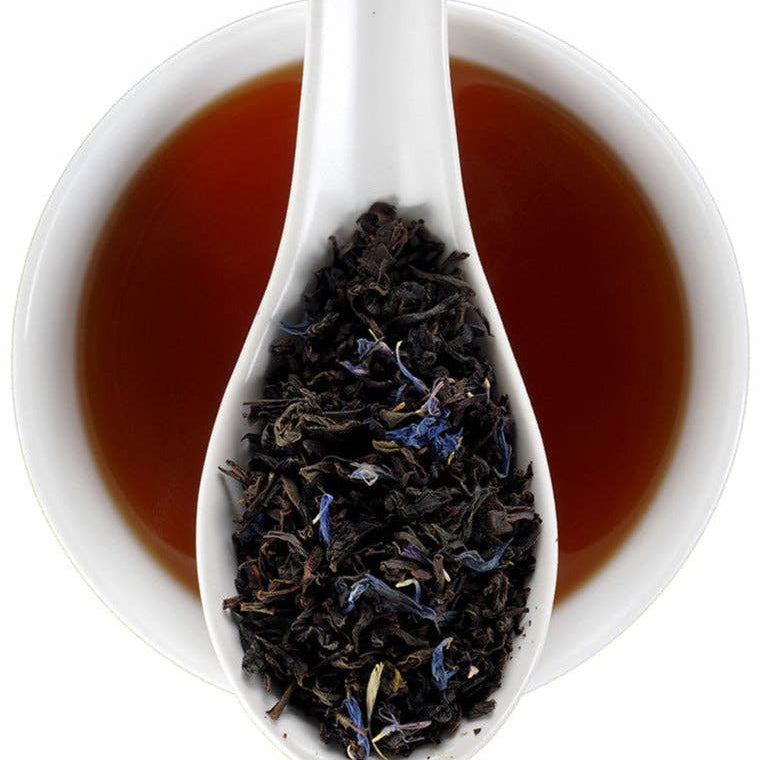 A white ceramic spoon filled with loose-leaf tea, set in front of a white bowl of amber-brown tea on a clean white background.