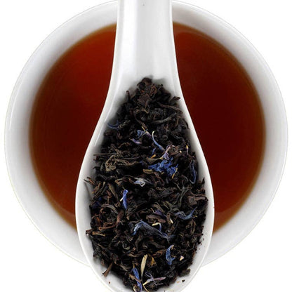 A white ceramic spoon filled with loose-leaf tea, set in front of a white bowl of amber-brown tea on a clean white background.