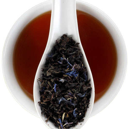 A white ceramic spoon filled with loose-leaf tea, set in front of a white bowl of amber-brown tea on a clean white background.