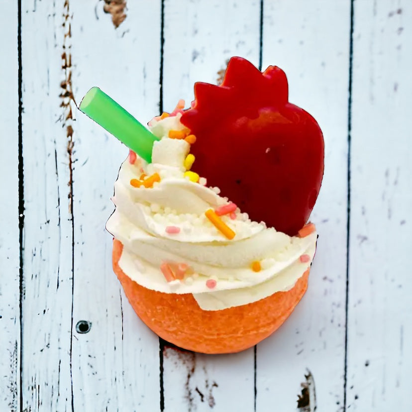 A round orange bath bomb with white frosting, colorful sprinkles, and a red orange strawberry soap/crayon on top against a wooden background