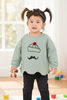Child wearing a light green sweater with a cake and mustache design, standing in a room with toys on the floor.