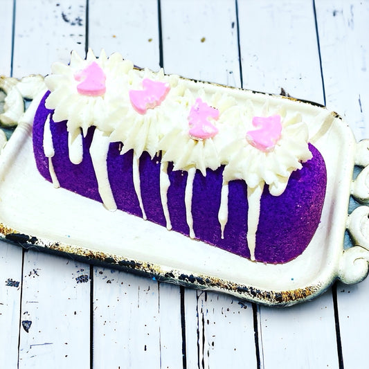 A purple twinkie bath bomb topped with white frosting and light purple sugar sprinkles on a white tray against a wood background