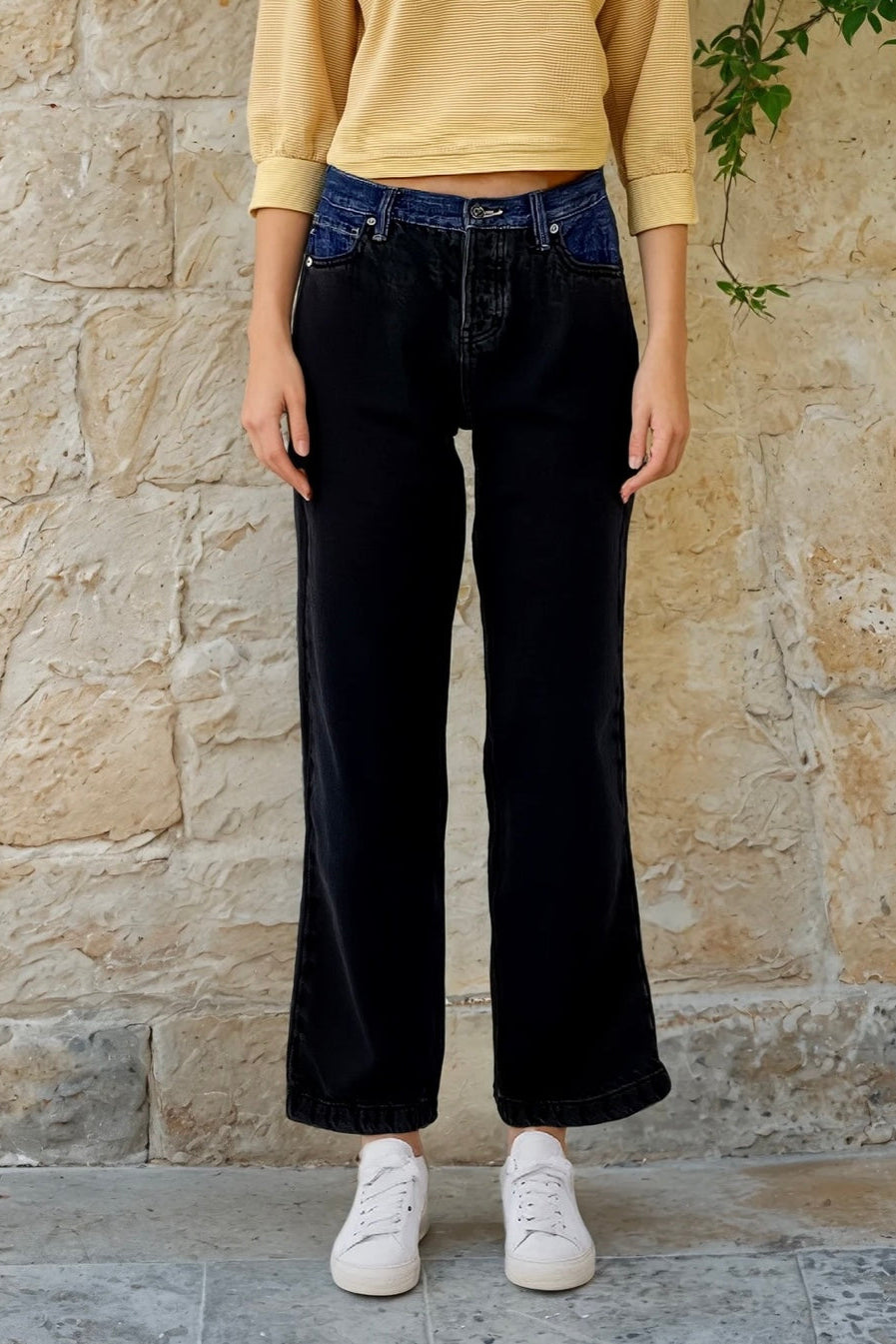 Person wearing black wide-leg jeans and a yellow top against a stone wall.